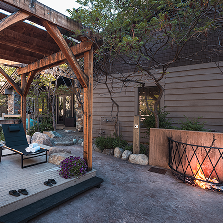Outdoor patio area with wooden pergola, lounge chair, fire pit, and landscaped garden beside a modern rustic building.