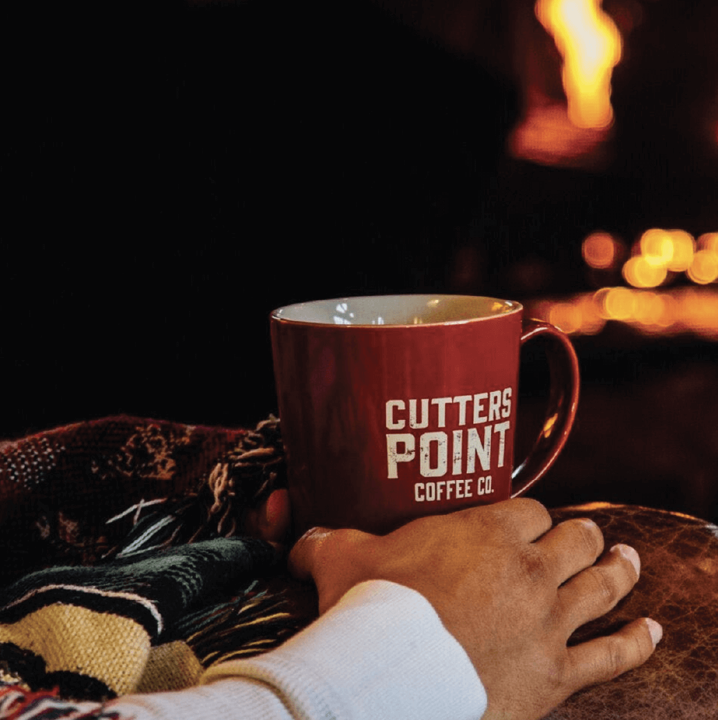A person holds a red Cutters Point Coffee Co. mug while sitting near a fireplace, with a blanket partially visible on their lap.