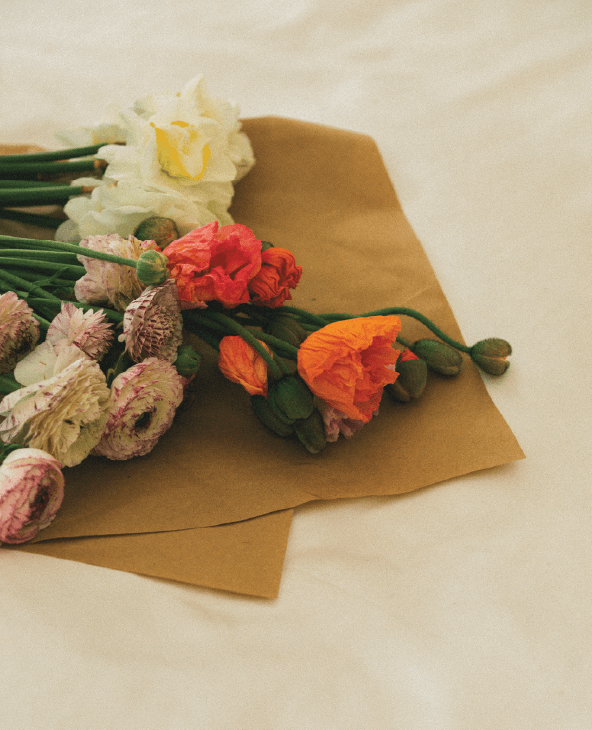 A bouquet of assorted flowers, including white, pink, and orange blooms, rests on a brown envelope atop a soft cream-colored surface.