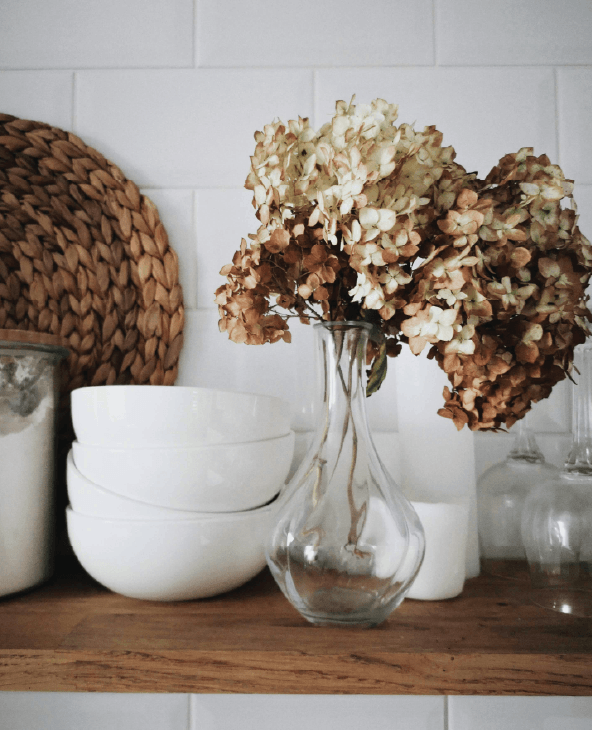A clear glass vase with dried hydrangeas sits on a wooden shelf next to stacked white bowls, a woven placemat, a glass jar, and white containers against a white tile wall.