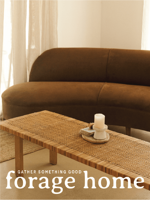 A cozy living room with a curved brown sofa, a woven wooden coffee table, a white candle, and neutral curtains. Text reads: Gather something good, forage home.