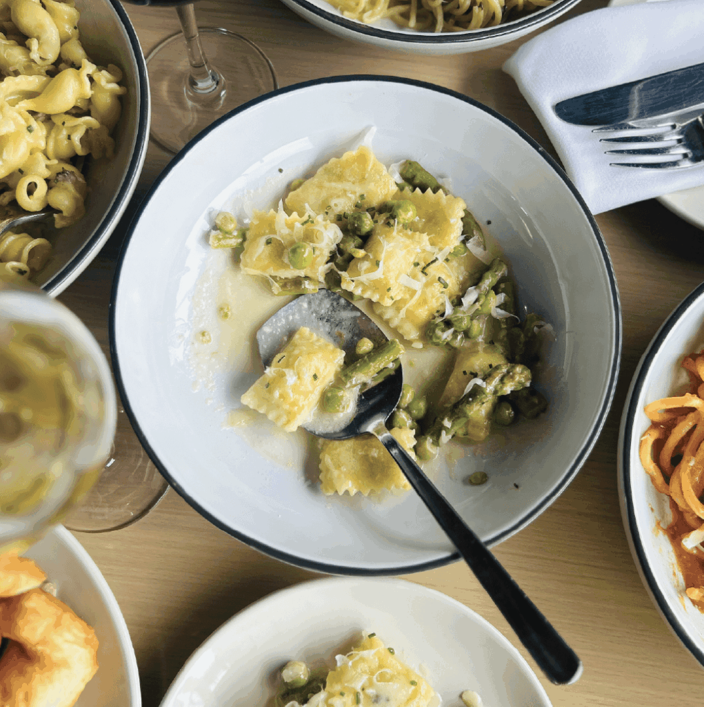 A plate of ravioli with asparagus and sauce is partially eaten, surrounded by other pasta dishes and a glass of white wine on a wooden table.