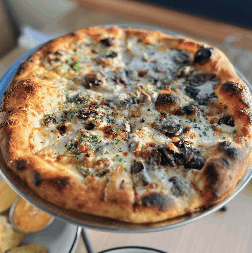 A freshly baked pizza topped with mushrooms, cheese, and herbs sits on a metal stand, with a lightly browned crust and some charred areas.