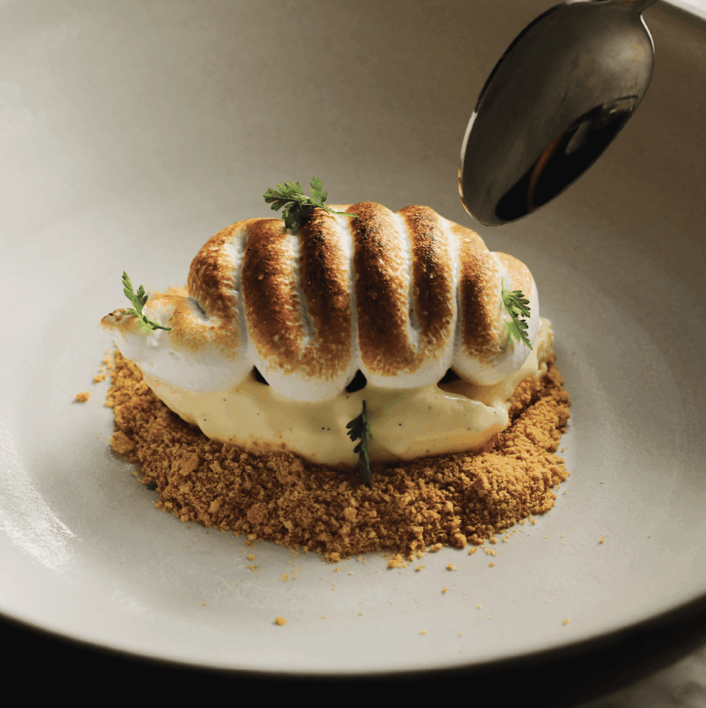 A plated dessert with toasted meringue, creamy filling, and crumble base, garnished with small green herbs, as a spoon is held above it.