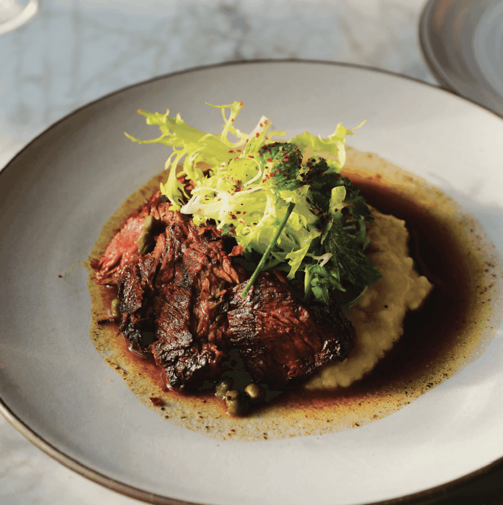 Plate of sliced grilled steak on mashed potatoes, topped with fresh greens and surrounded by a brown sauce.
