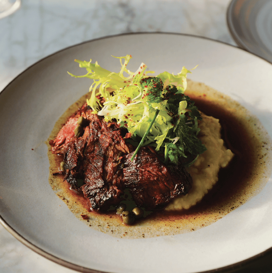 Plate of sliced grilled steak on mashed potatoes, topped with fresh greens and surrounded by a brown sauce.
