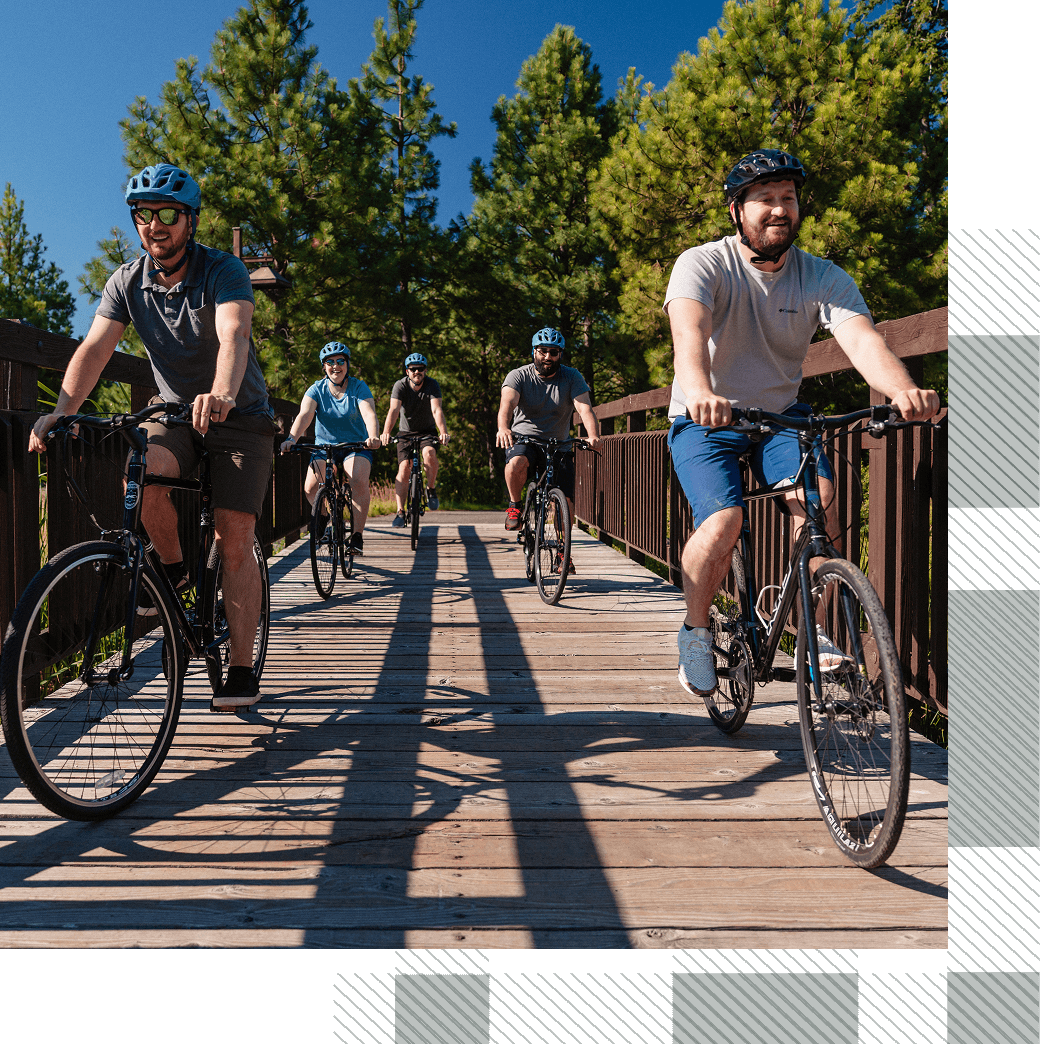 Five adults ride bicycles across a wooden bridge surrounded by trees on a sunny day.