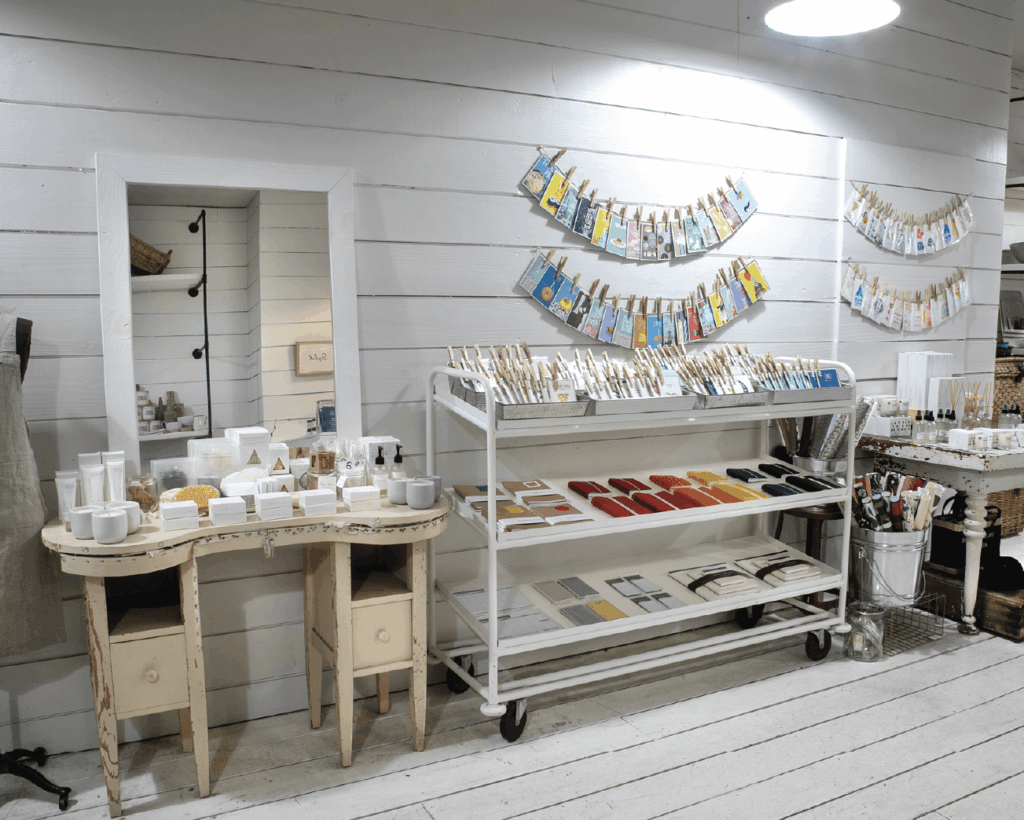 A boutique display features various soaps, bath products, and stationery on shelves and a vintage vanity in a white wooden room with organized, colorful packaging.
