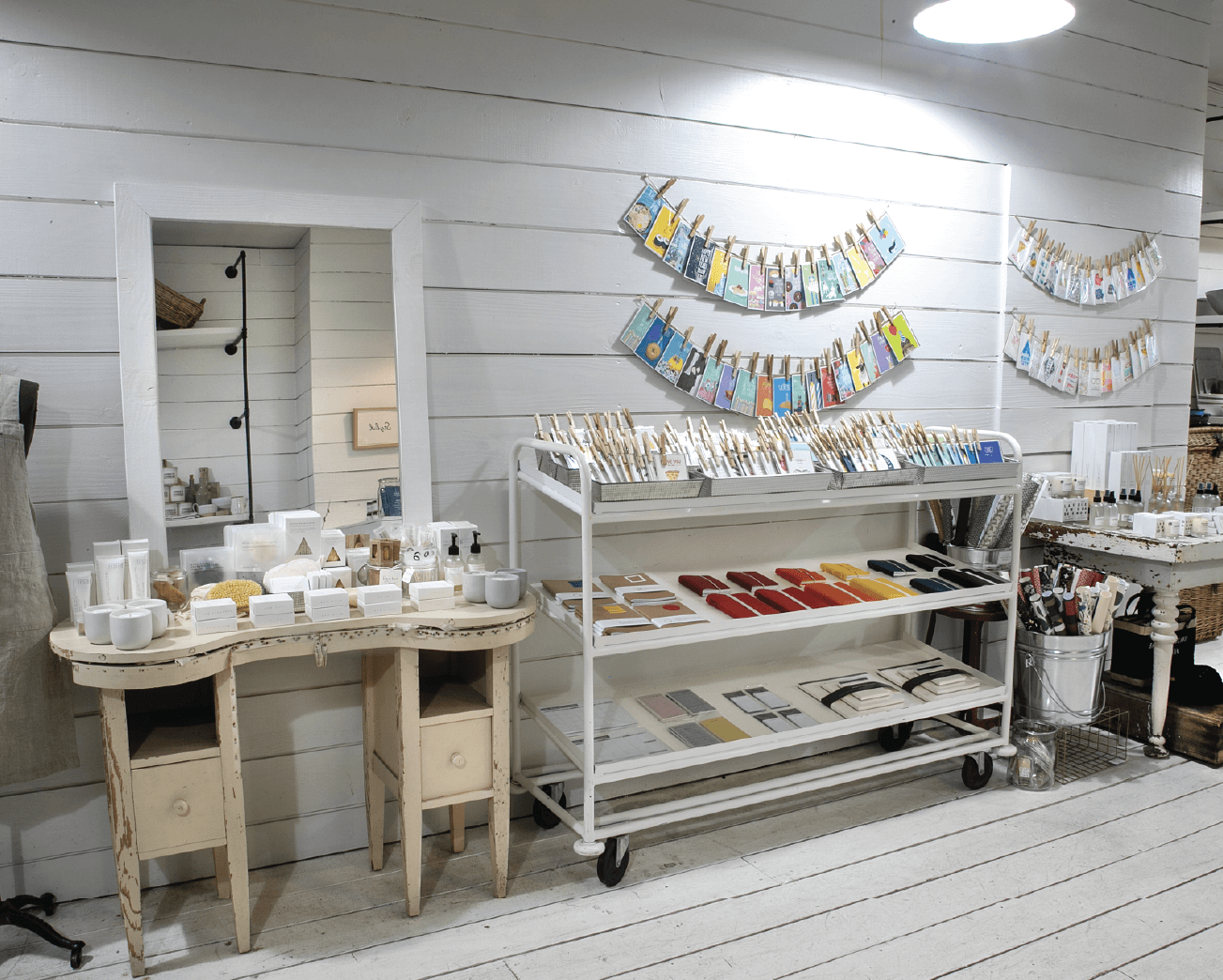 A boutique display features various soaps, bath products, and stationery on shelves and a vintage vanity in a white wooden room with organized, colorful packaging.
