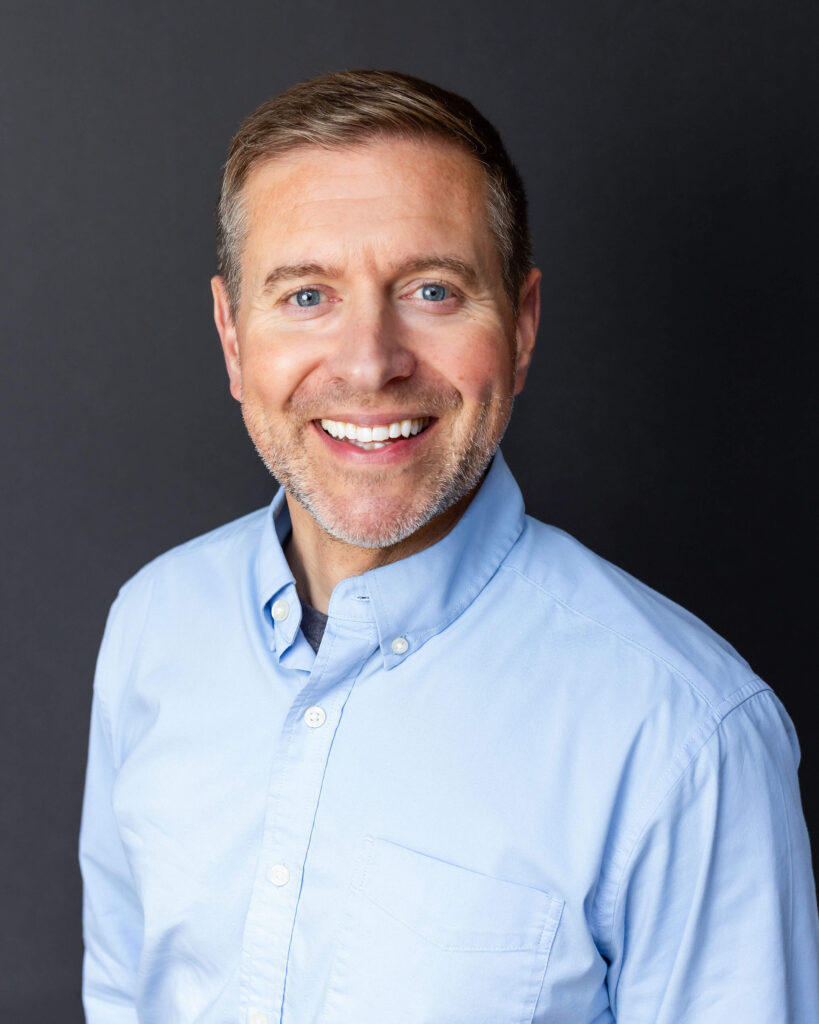 A man with short brown hair and a trimmed beard smiles while wearing a light blue button-up shirt against a plain dark background.
