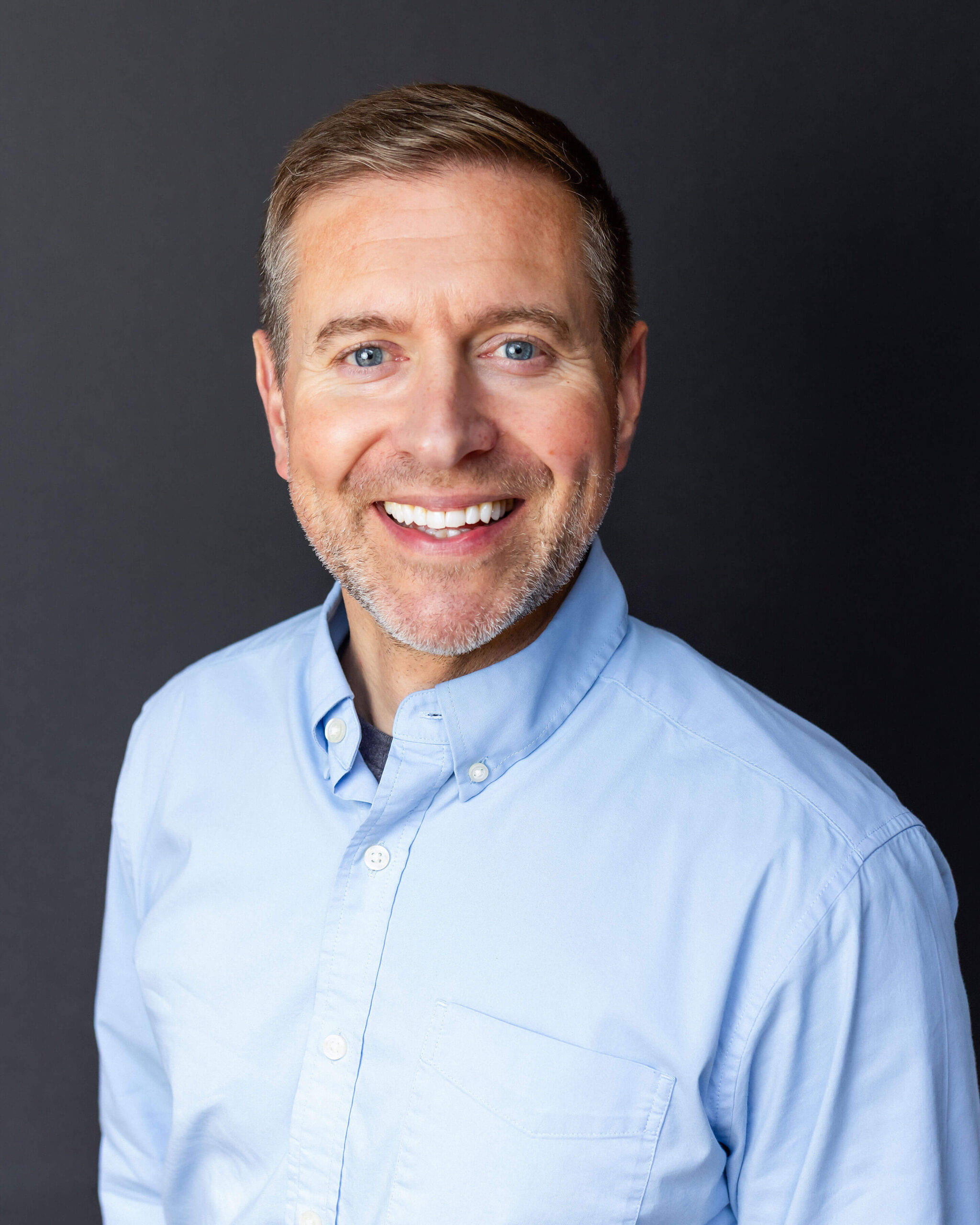 A man with short brown hair and a trimmed beard smiles while wearing a light blue button-up shirt against a plain dark background.