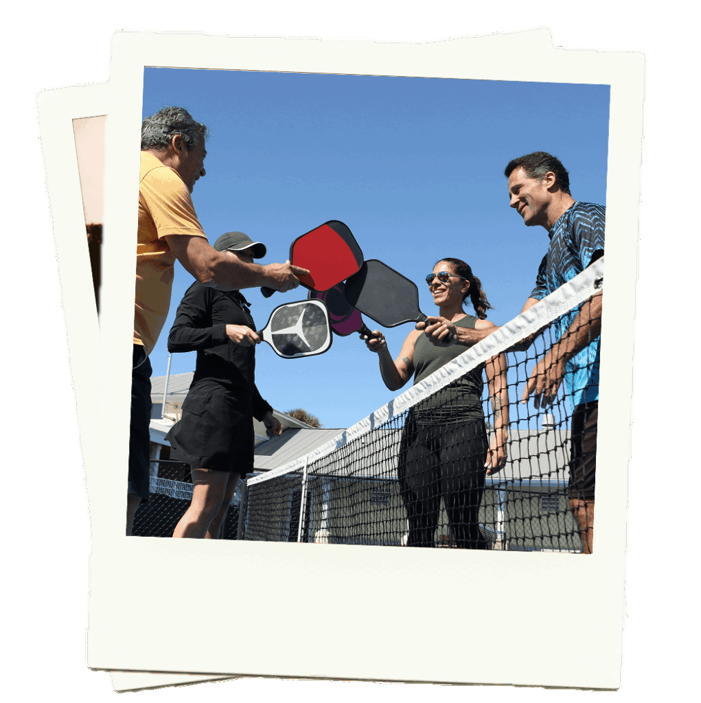 Four adults stand by a pickleball net outdoors, each holding a paddle and smiling as they greet or congratulate each other.