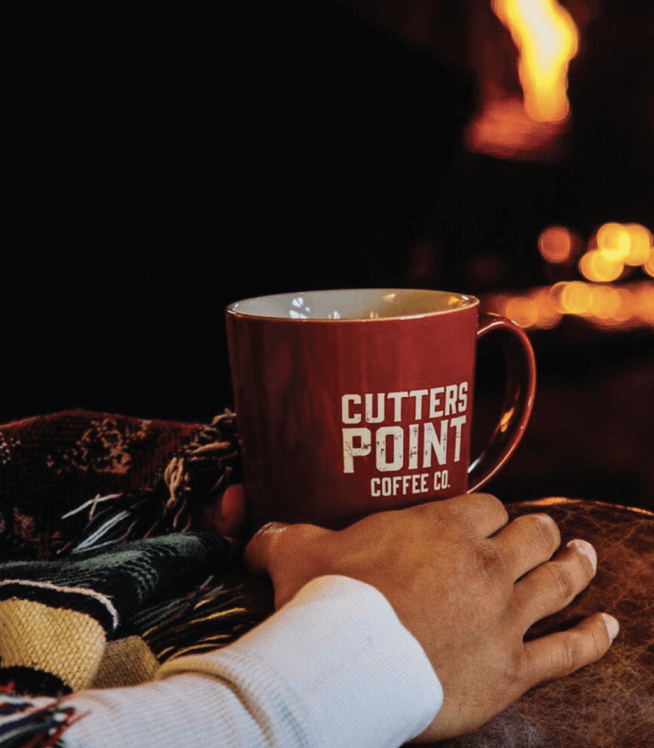 A hand rests on a table next to a red Cutters Point Coffee Co. mug, with a blanket and a fireplace glowing in the background.