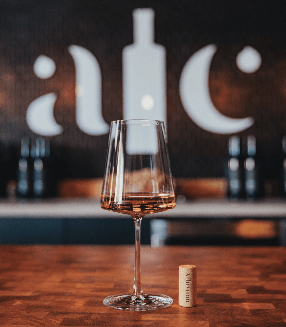 A wine glass with white wine sits on a wooden counter next to a cork, with a blurred background featuring large, stylized letters and bottle shapes on the wall.