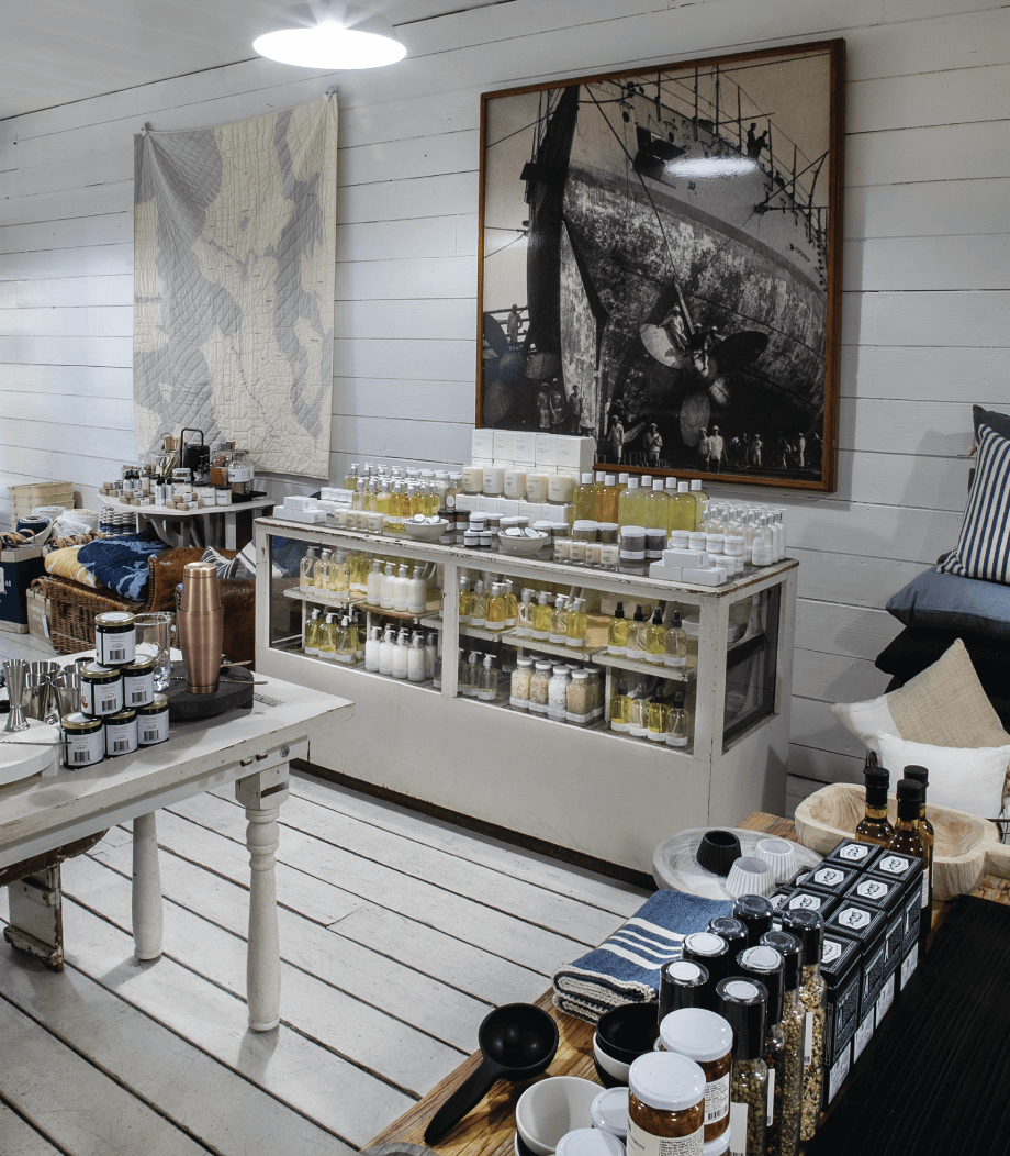 A shop interior with white wooden walls and floors, displaying shelves of bottles, jars, and home goods, with framed nautical artwork on the walls.