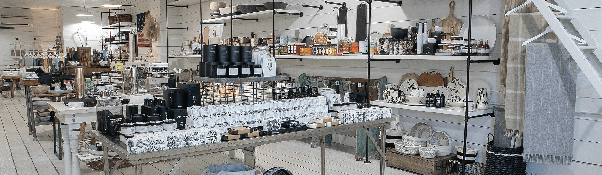 A home goods store interior with shelves and tables displaying candles, jars, kitchenware, utensils, and decorative items in a neutral color palette.
