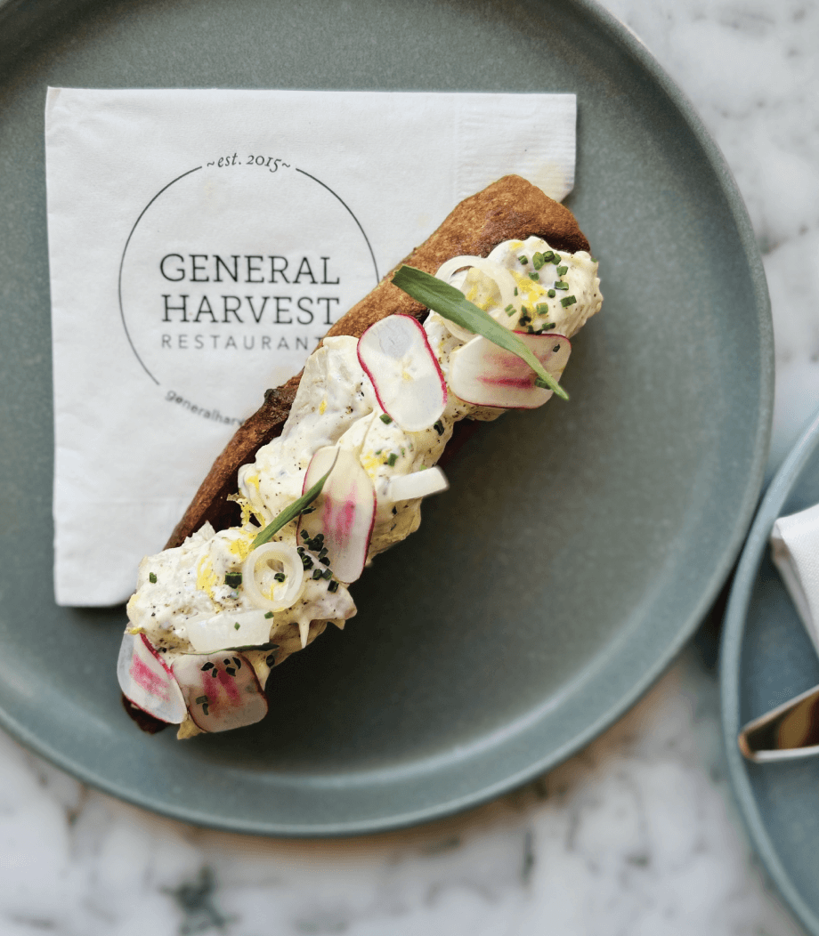 An open-faced sandwich topped with sliced radishes, greens, and cream on a blue plate, placed on a napkin with the General Harvest Restaurant logo.