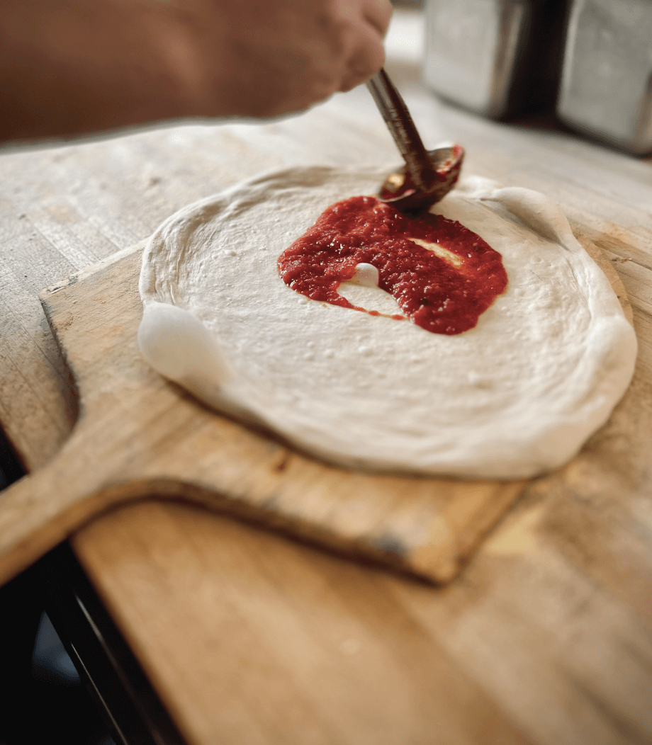 A hand spreads tomato sauce onto a circular pizza dough placed on a wooden pizza peel.