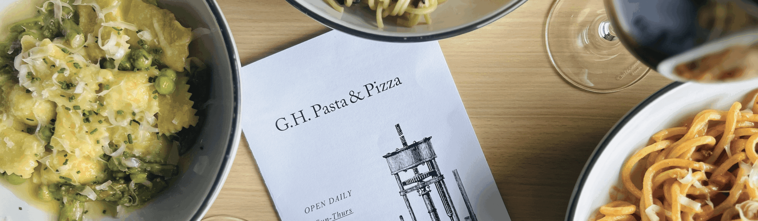 Overhead view of pasta dishes, a menu labeled "G.H. Pasta & Pizza," and a glass of red wine on a wooden table.
