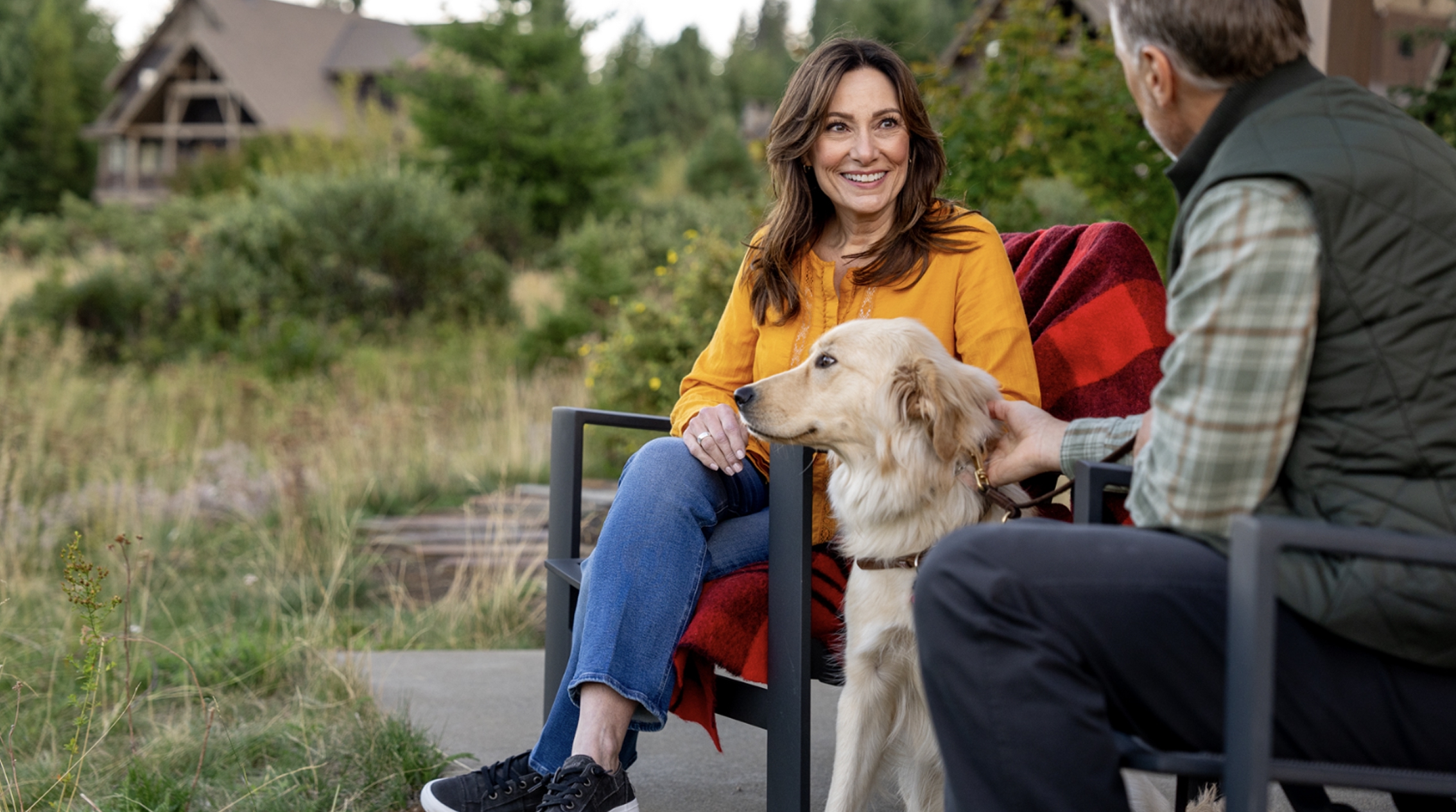 Two people sit outdoors on patio chairs, talking, with a golden dog beside them. Trees and a house are visible in the background.