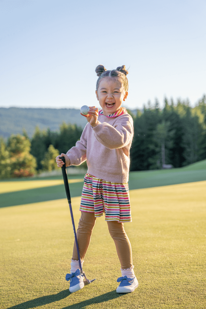 Young child smiling on a golf course, holding a golf club and showing a golf ball, with trees and hills in the background.