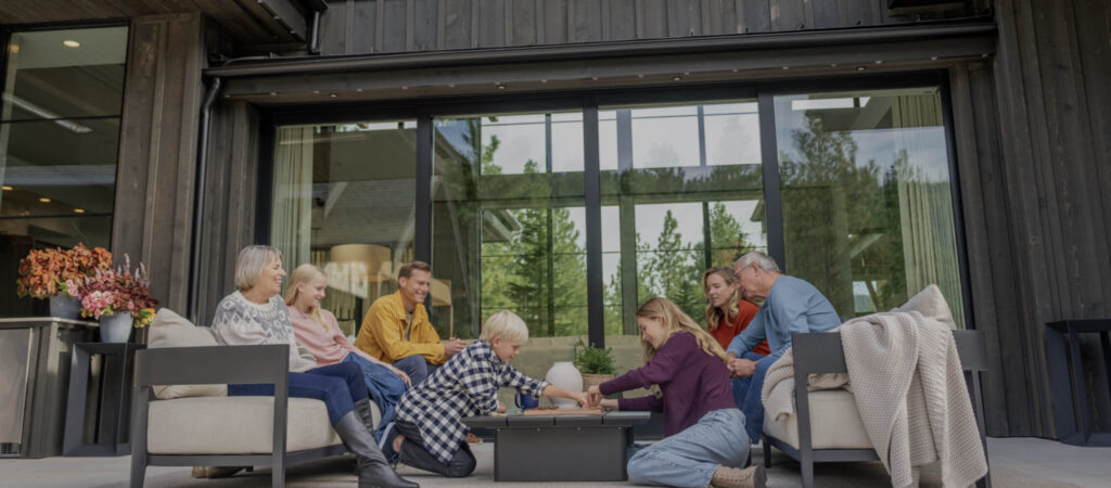 A family of seven, including adults and children, sits around a table on a patio outside a modern house, playing a game together on a sunny day.