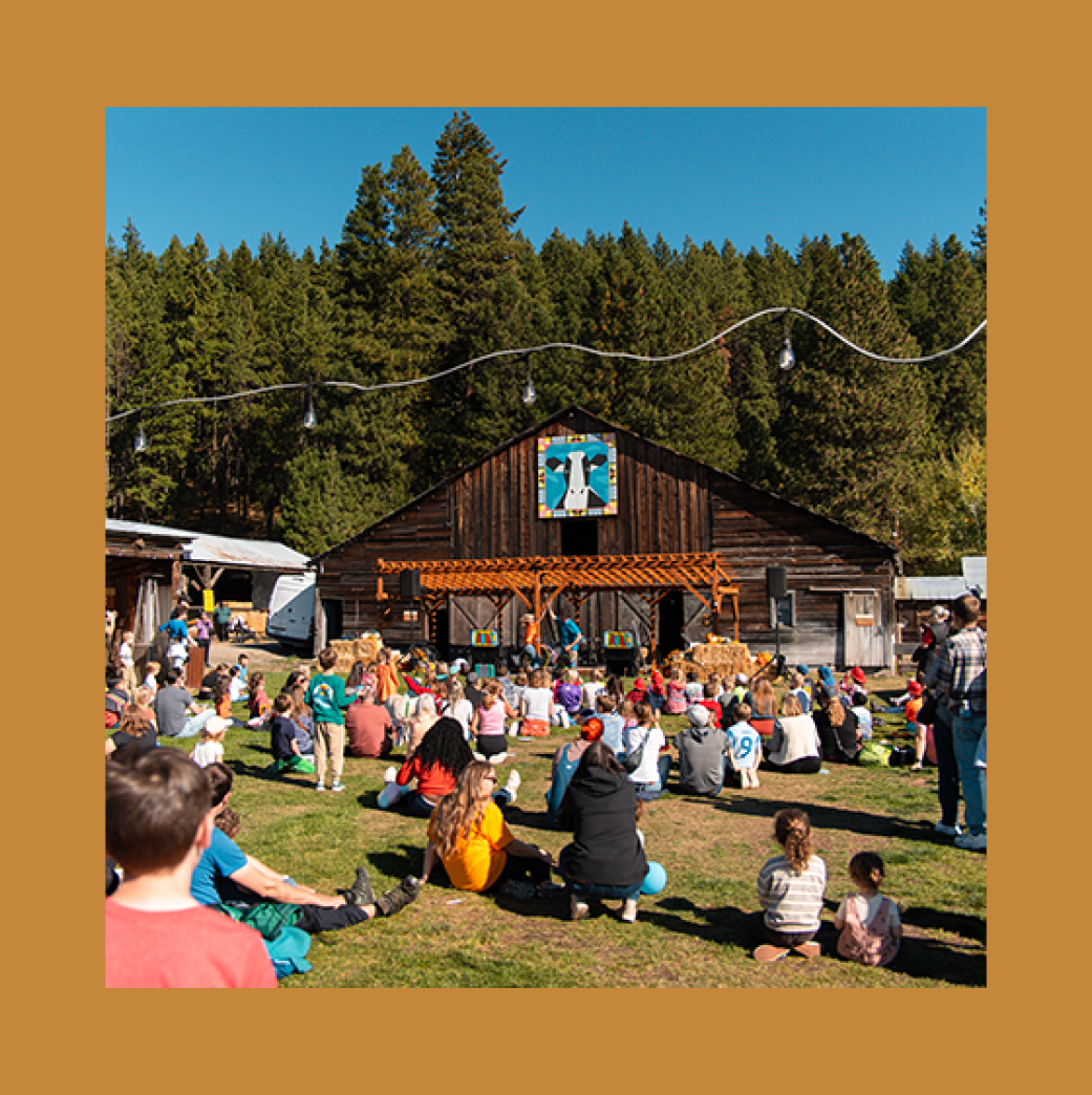 A crowd of people sits on grass facing a rustic wooden stage in front of a barn, surrounded by trees under a clear blue sky.