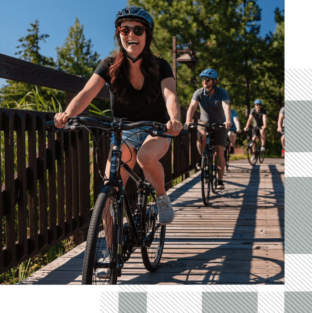 A group of people wearing helmets ride bicycles across a wooden bridge on a sunny day, with trees and greenery in the background.