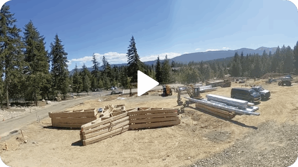 Construction site with stacked lumber, a forklift, a black van, and metal beams on a dirt lot surrounded by trees and distant mountains under a clear sky.