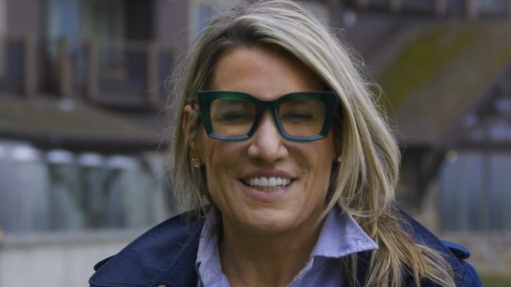 A woman with long blonde hair, wearing large dark-framed glasses and a blue jacket, stands outdoors in front of a blurred building.