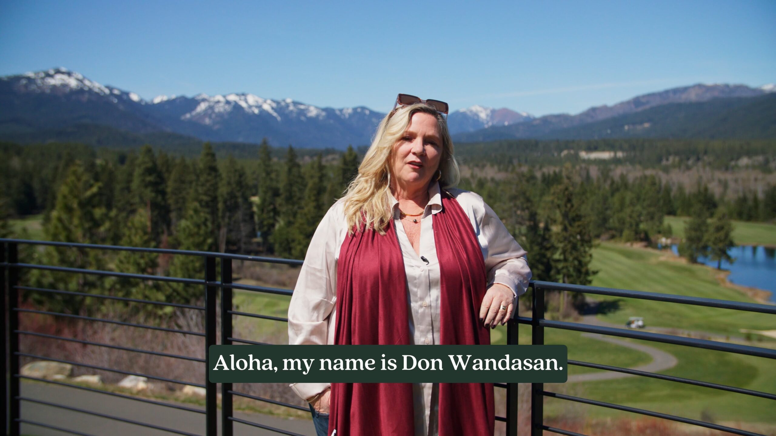 A woman stands on a balcony overlooking a scenic landscape with trees, a golf course, and snow-capped mountains; she wears a white blouse and red scarf—an idyllic morning for Dawn Wandasan of Suncadia Real Estate.