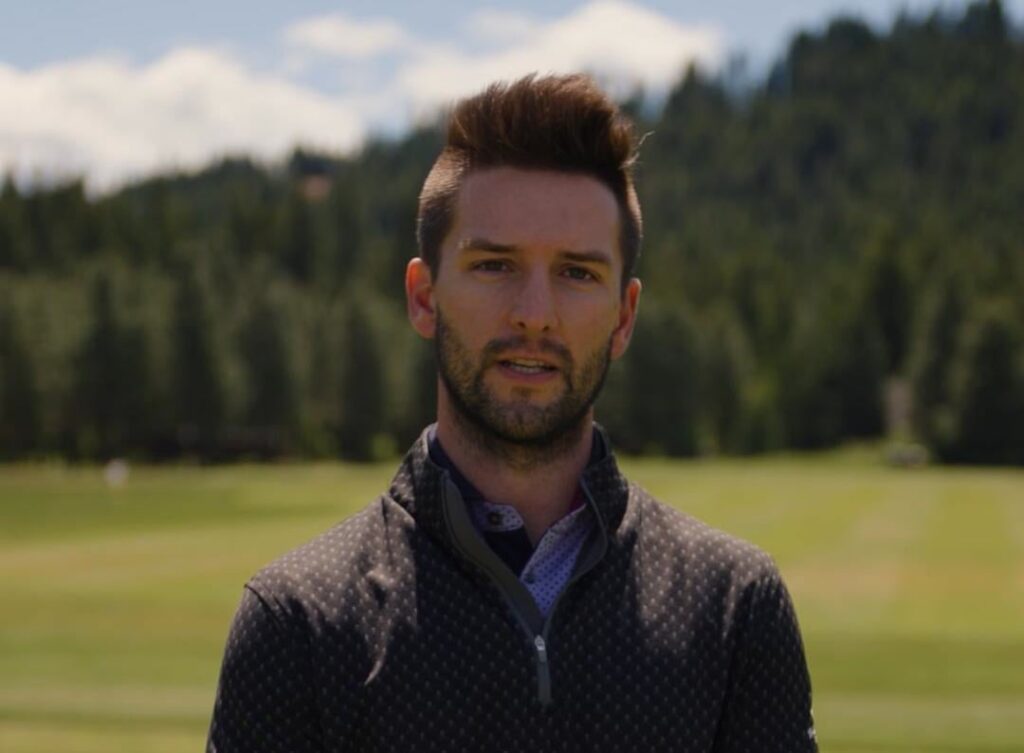 A man with short brown hair and a trimmed beard stands outdoors on a grassy field with trees and hills in the background.