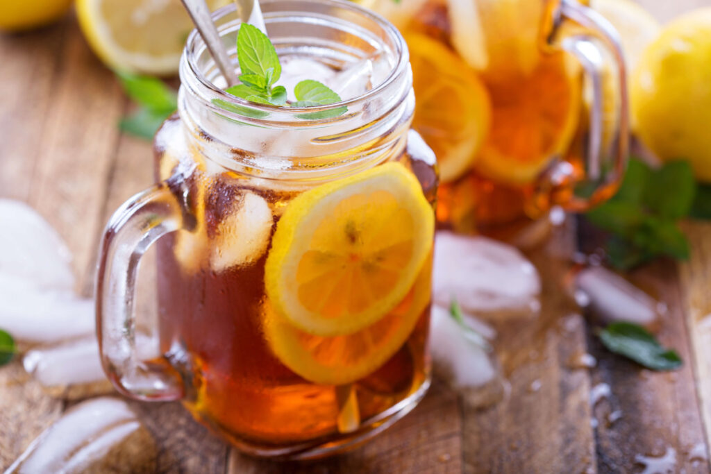 A mason jar filled with iced tea, lemon slices, ice cubes, and a mint sprig, placed on a wooden surface with scattered ice and a lemon in the background.