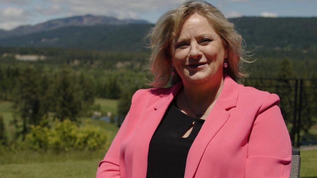 A woman wearing a bright pink blazer sits outdoors with a scenic background of trees, hills, and a partly cloudy sky.