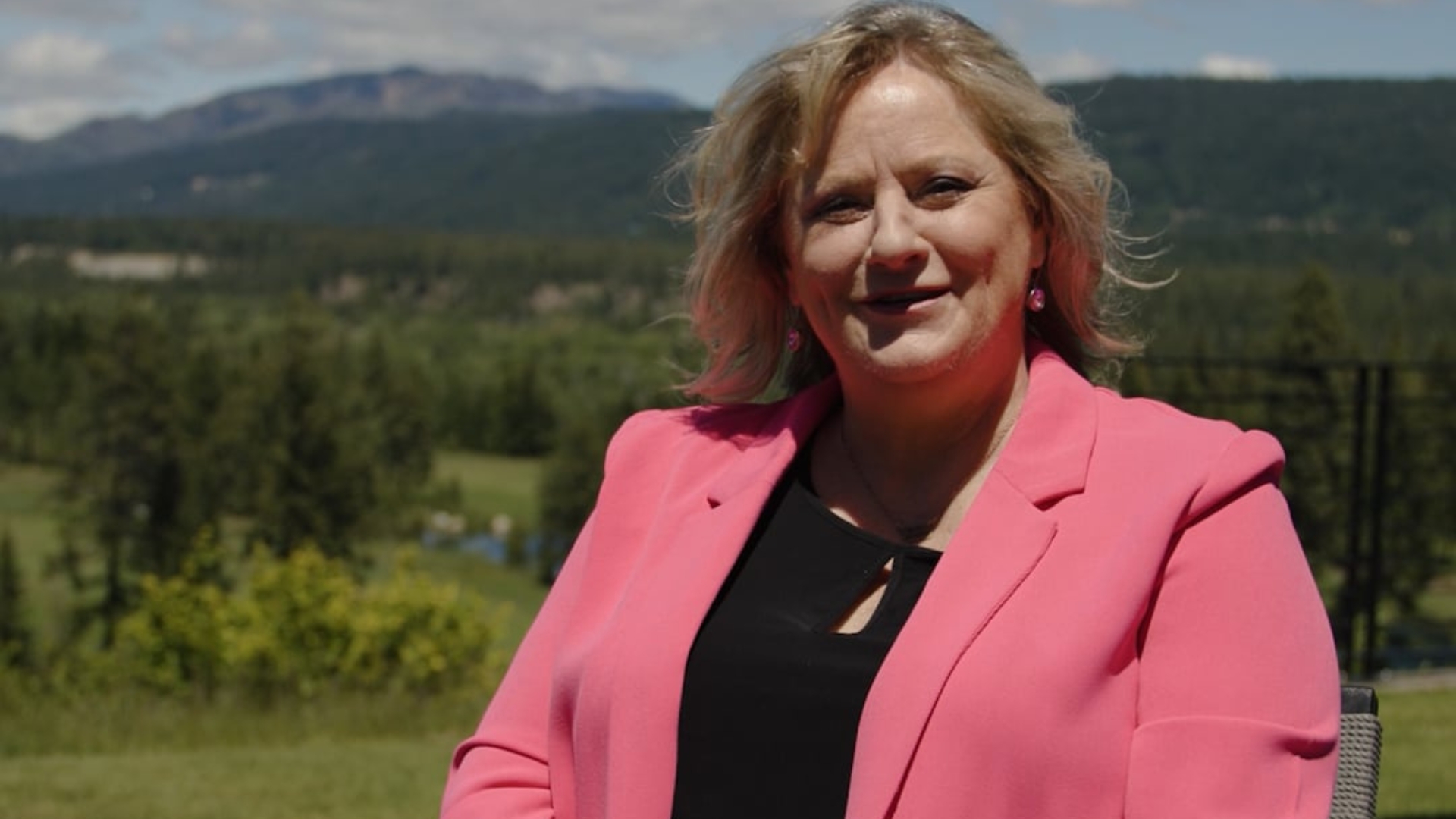 A woman wearing a bright pink blazer sits outdoors with a scenic background of trees, hills, and a partly cloudy sky.