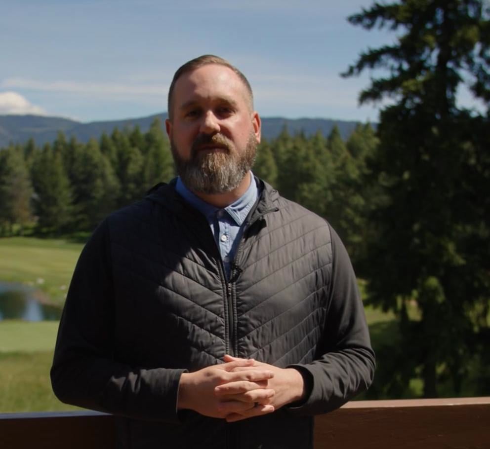 A man with a beard stands outdoors on a wooden deck, with a golf course, trees, and mountains visible in the background.