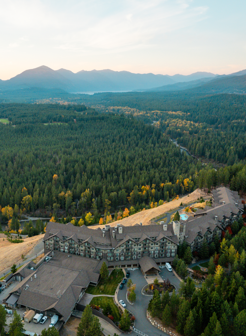 A large lodge-style building sits on a hillside surrounded by dense forest, with mountains visible in the distance under a partly cloudy sky.