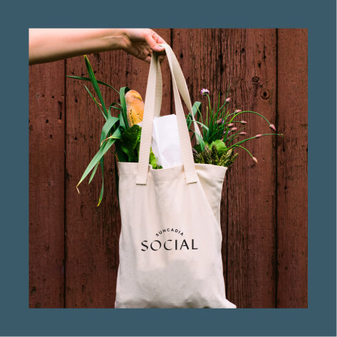 A hand holds a tote bag filled with groceries and flowers against a wooden background. The bag has the word "SOCIAL" printed on it.