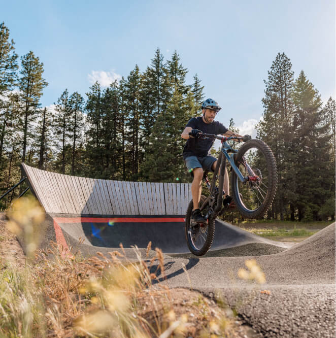 A person wearing a helmet rides a bicycle over a paved bike park feature, lifting the front wheel off the ground, with trees in the background.
