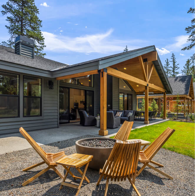 A modern house with a covered patio, outdoor seating area, four wooden chairs around a fire pit, and a green lawn surrounded by trees on a sunny day.