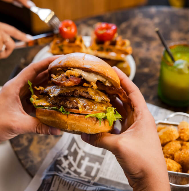 A person holds a large cheeseburger with bacon, lettuce, and sauce at a table set with tater tots, a drink, and a plate of waffle fries topped with tomatoes.