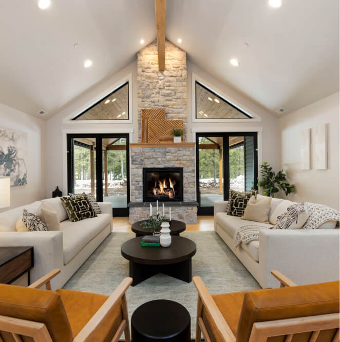 Modern living room with two white sofas, wooden chairs, a round coffee table, and a stone fireplace beneath a vaulted ceiling with exposed wood beam. Large windows show outdoor trees.