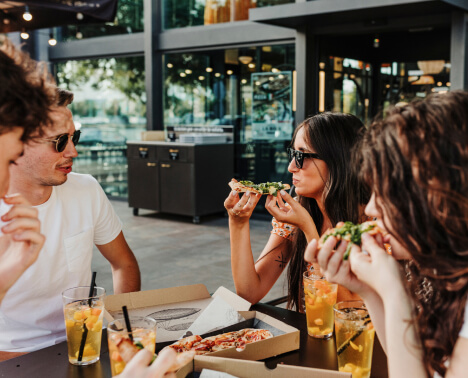 Four people sit at an outdoor table eating pizza and drinking beverages with ice, with a modern glass building in the background.