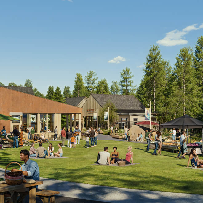 People gather on a grassy lawn outside modern wooden buildings on a sunny day, sitting, eating, and socializing amid trees and market stalls.