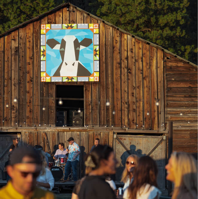 A band performs on a stage inside a rustic wooden barn with a geometric cow mural above, while people gather in the foreground.