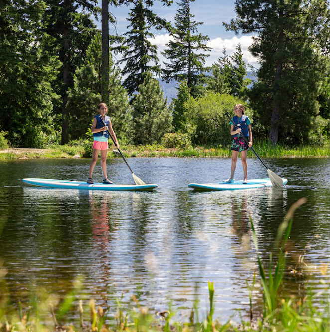 Two people wearing life jackets paddle standing on paddleboards on a calm lake surrounded by trees and greenery.