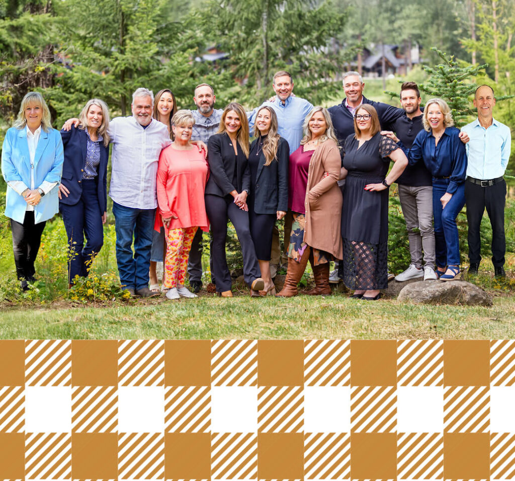 A group of fourteen adults pose together outdoors in front of trees, standing on grass, with a brown and white checkered pattern at the bottom of the image.