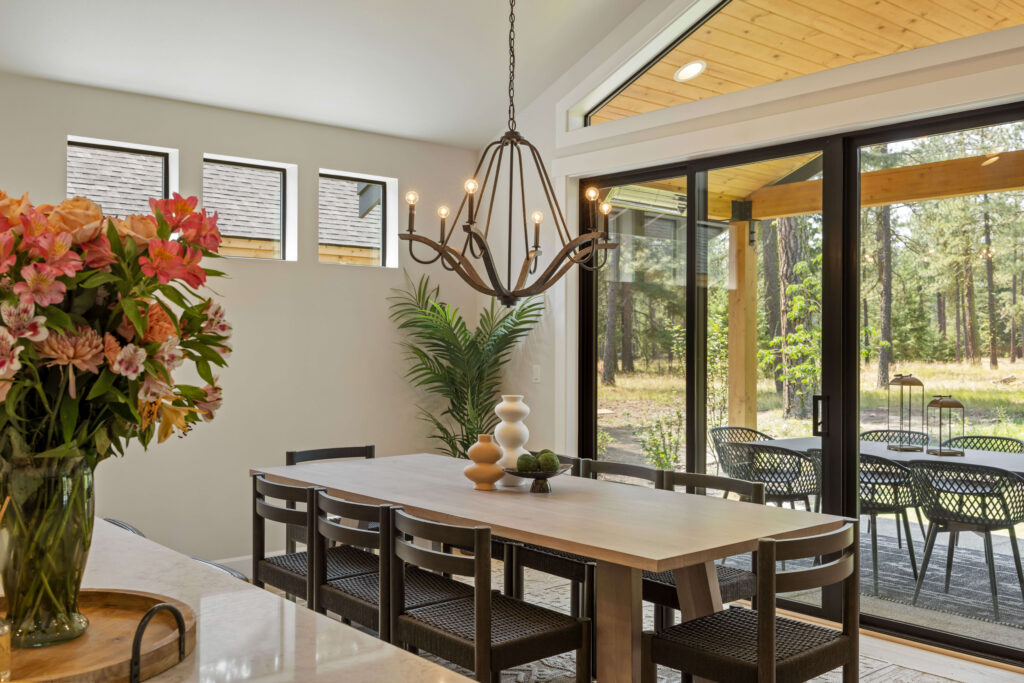 Modern dining room with a wooden table, black chairs, a floral centerpiece, and a chandelier. Large windows and glass doors overlook a patio and wooded outdoor area.