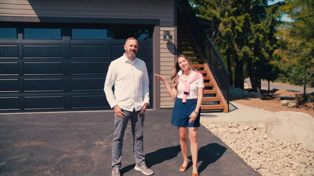 A man and a woman stand outside on a driveway in front of a garage with stairs leading up to a deck, surrounded by trees and rocks.