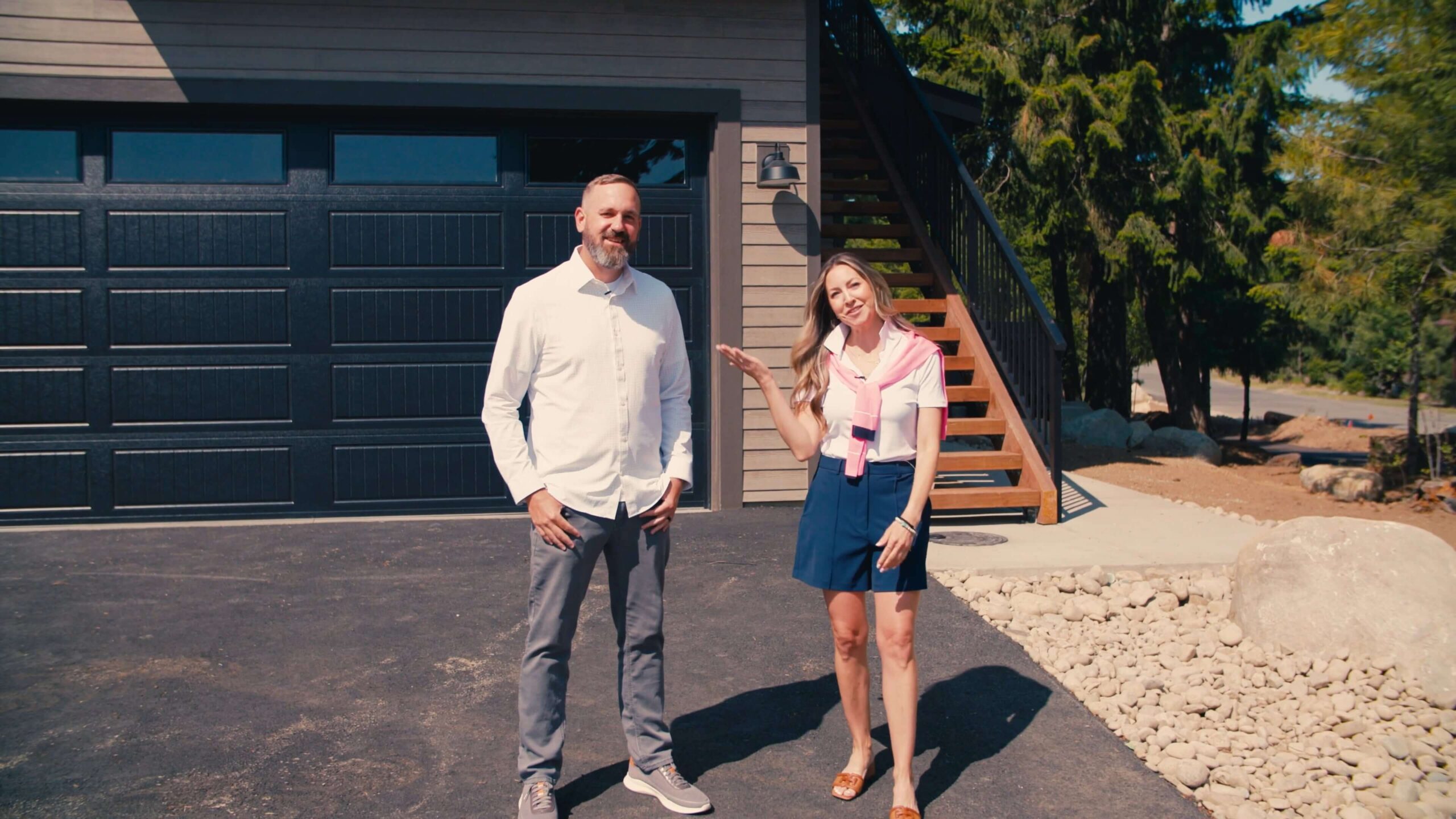 A man and a woman stand outside on a driveway in front of a garage with stairs leading up to a deck, surrounded by trees and rocks.